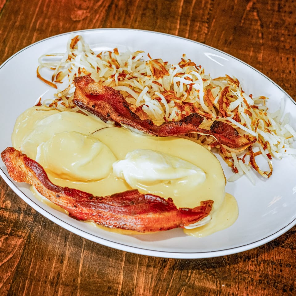 Eggs Benedict with hollandaise sauce, crispy bacon, and hash browns on a white plate.