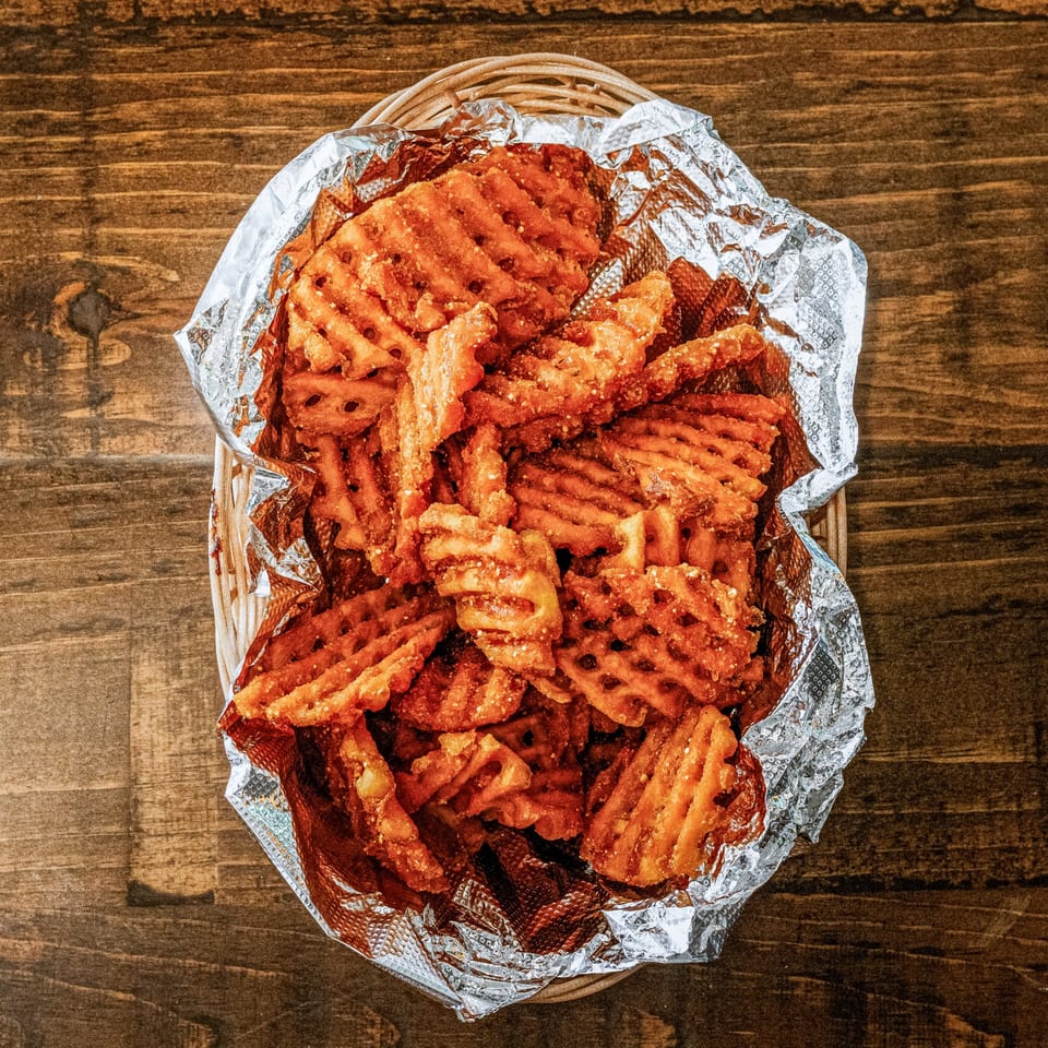 A basket of crispy, golden-brown waffle fries, seasoned with salt, served in foil on a wooden table.