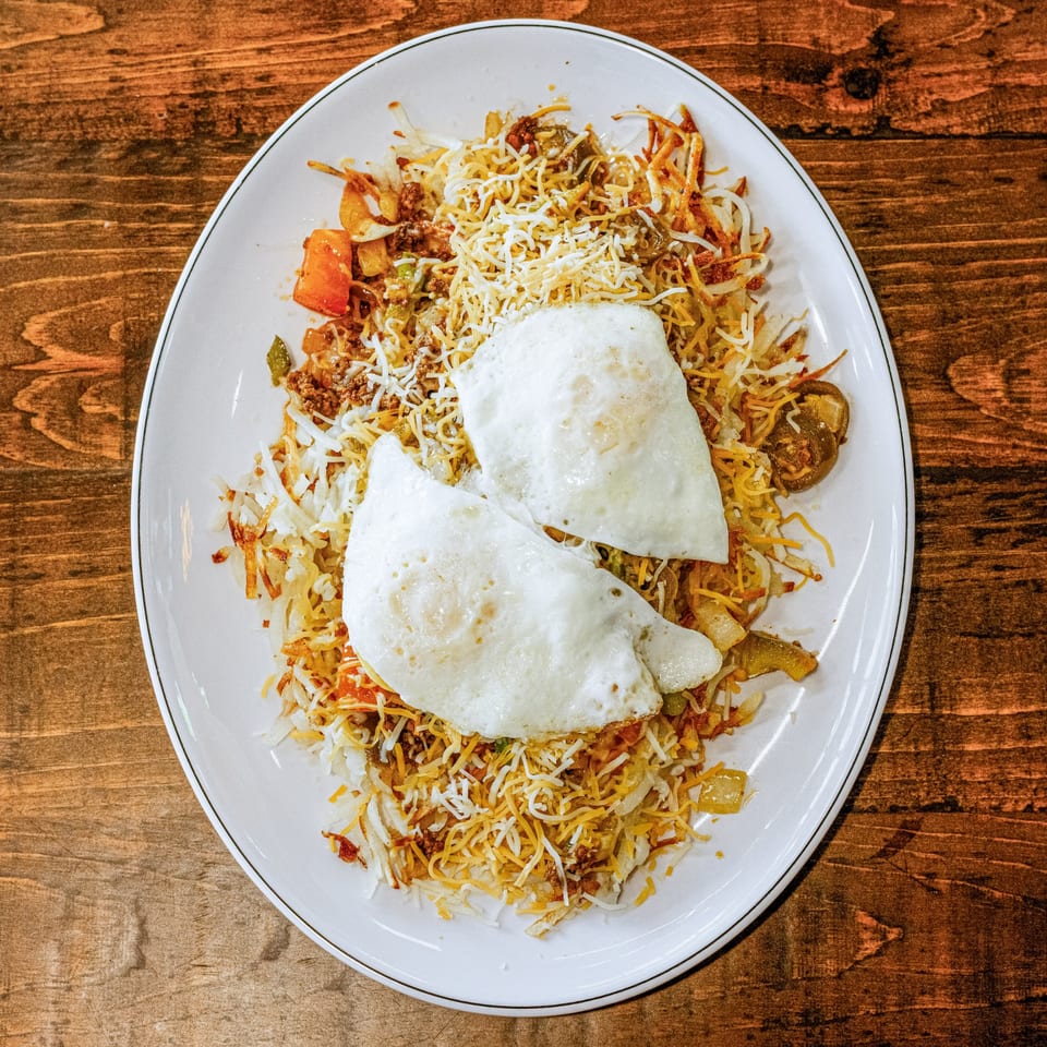 A diner breakfast skillet with hash browns, ground meat, vegetables, shredded cheese, and two fried eggs.