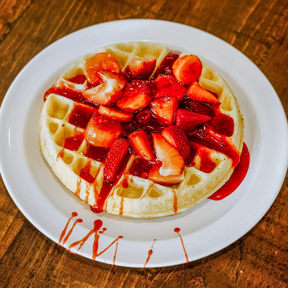 A golden waffle topped with fresh strawberries and strawberry syrup on a white plate at HWY 47 KITCHEN.