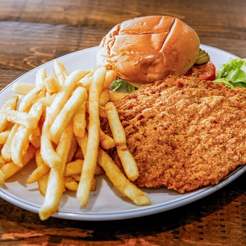 A breaded pork tenderloin sandwich with fries, lettuce, tomato, and pickles on a white plate.