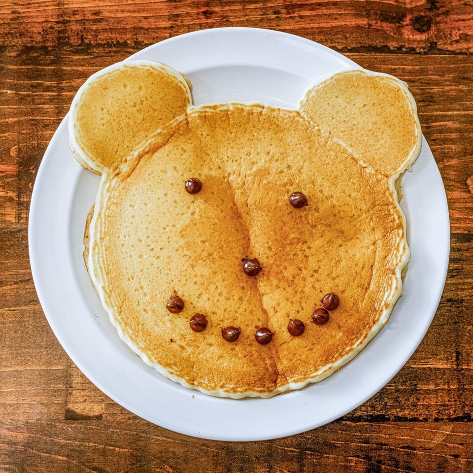 A bear-shaped pancake with chocolate chip eyes, nose, and mouth on a white plate at HWY 47 KITCHEN.