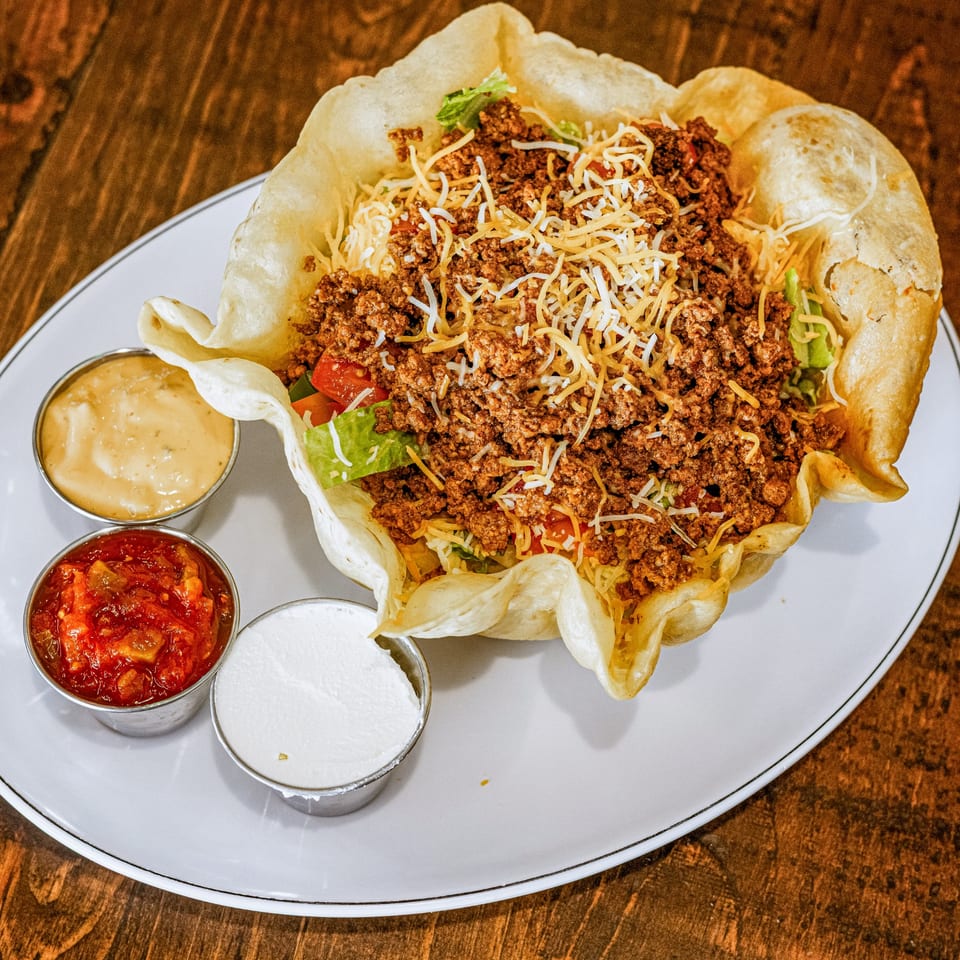Taco salad in a crispy tortilla bowl with ground beef, cheese, lettuce, and sides of salsa, queso, and sour cream.