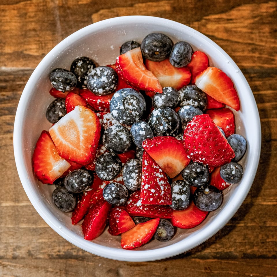 A white bowl filled with fresh strawberries and blueberries dusted with powdered sugar.
